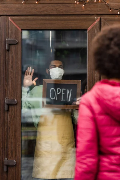 African american barista in medical mask waving hand and holding chalkboard with open lettering in cafe near client — Stock Photo