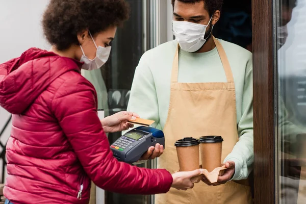 Barista afroamericano en máscara médica sosteniendo café y terminal de pago cerca del cliente con tarjeta de crédito - foto de stock
