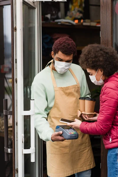 Barista em máscara médica segurando terminal de pagamento perto do cliente com smartphone e café no café — Fotografia de Stock