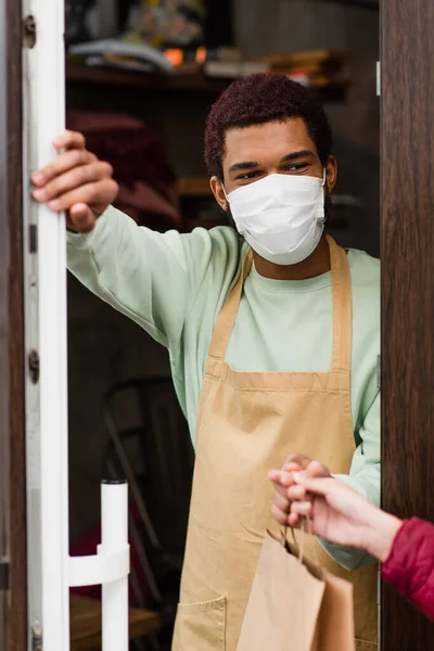 African american barista in medical mask giving paper bag to blurred client near door of cafe — Stock Photo