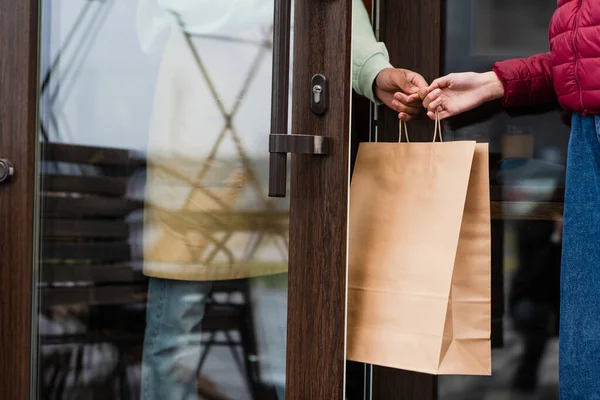 Cropped view of african american barista giving paper bag to client near door of cafe — Stock Photo