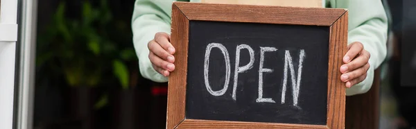 Cropped view of african american barista holding chalkboard with open lettering near entrance of cafe, banner — Stock Photo