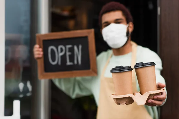 Café para llevar en la mano de barista afroamericano en máscara médica sosteniendo pizarra con letras abiertas sobre fondo borroso - foto de stock