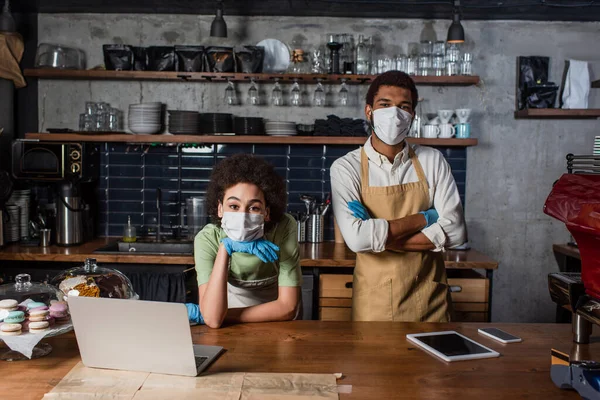 Африканський барист у медичній масці та рукавичках з латексу стоїть біля приладів та колег у кафе. — стокове фото