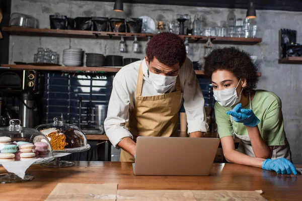 African american barista in medical mask pointing at laptop near colleague in cafe — Stock Photo