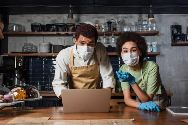 Baristas afroamericanos en máscaras médicas y guantes de látex mirando la cámara cerca de la computadora portátil en la cafetería - foto de stock