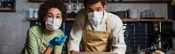 Baristas afro-americanos em máscaras médicas olhando para a câmera no café, bandeira — Fotografia de Stock