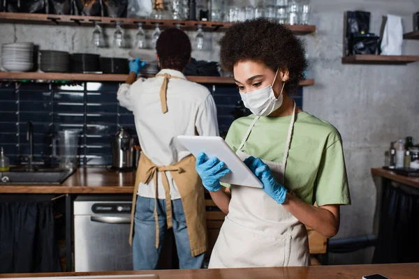African american barista in medical mask and latex gloves using digital tablet near blurred colleague — Stock Photo