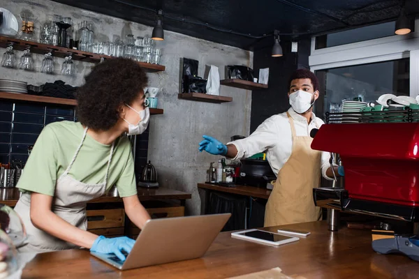 Africano americano barista em máscara médica de pé perto colega turvo usando laptop no café — Fotografia de Stock