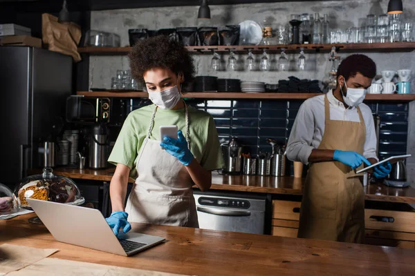 African american barista in medical mask using smartphone and laptop near colleague in cafe — Stock Photo