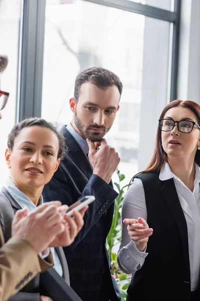 Empresaria apuntando a colega borroso con teléfono inteligente cerca de gente de negocios multiétnicos - foto de stock