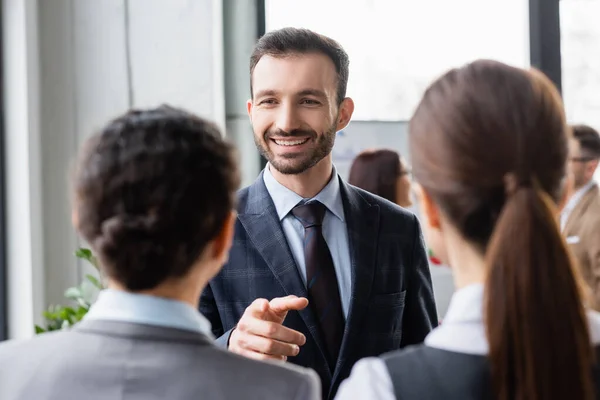 Hombre de negocios sonriente señalando con el dedo cerca de colegas borrosos en la oficina - foto de stock