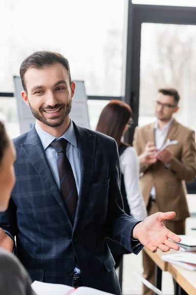 Empresário sorridente conversando com colega desfocado com caderno no escritório — Fotografia de Stock