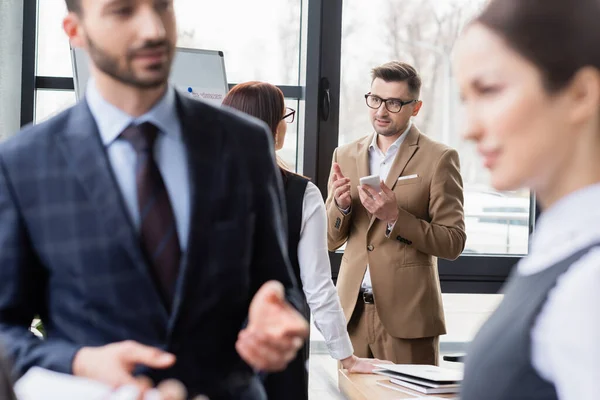 Businessman with smartphone pointing with finger near colleague in office — Stock Photo