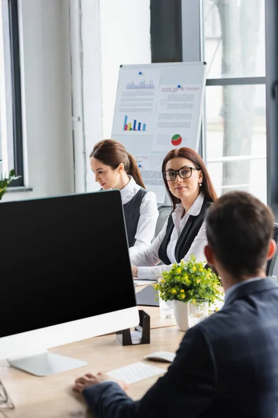 Donna d'affari che guarda la fotocamera vicino a collega offuscata e computer con schermo vuoto in ufficio — Foto stock