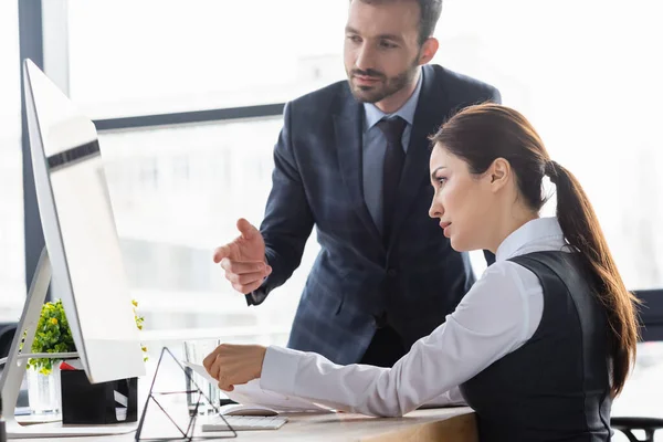 Geschäftsfrau hält Dokumente neben Kollegin und zeigt auf Computer im Büro — Stockfoto