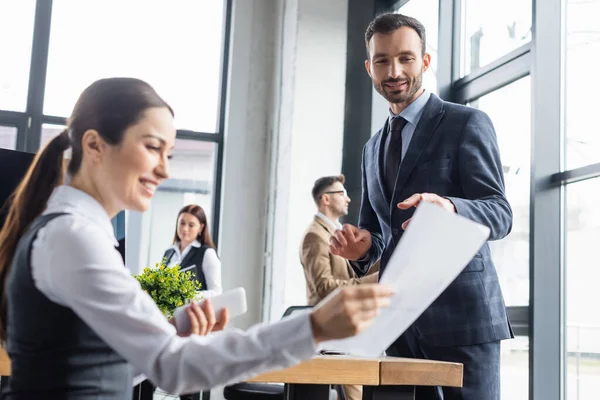 Alegre hombre de negocios de pie cerca borrosa colega con teléfono inteligente y papel en la oficina - foto de stock