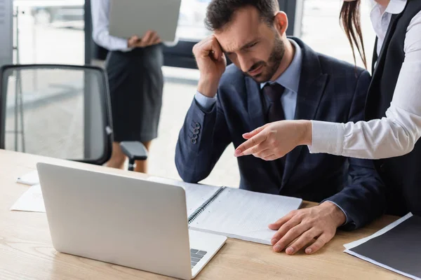 Femme d'affaires pointant vers ordinateur portable près bouleversé homme d'affaires et dossier en papier dans le bureau — Photo de stock