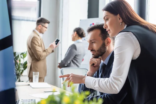 Vista lateral de la mujer de negocios apuntando a la computadora portátil cerca de hombre de negocios con pluma en la oficina — Stock Photo