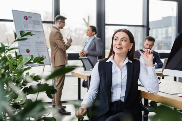 Smiling businesswoman with pen sitting near computer and plant in office — Stock Photo