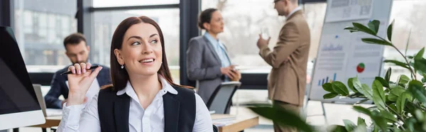 Mujer de negocios con pluma sonriendo mientras trabaja cerca de la computadora y colegas borrosos, pancarta - foto de stock