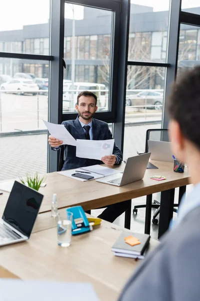 Businessman holding documents near blurred colleague and laptops in office — Stock Photo