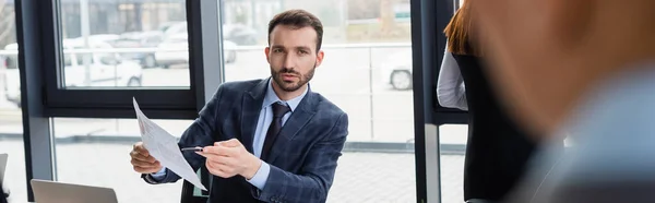 Uomo d'affari in giacca e cravatta con carta e penna vicino a collega sfocato, striscione — Foto stock