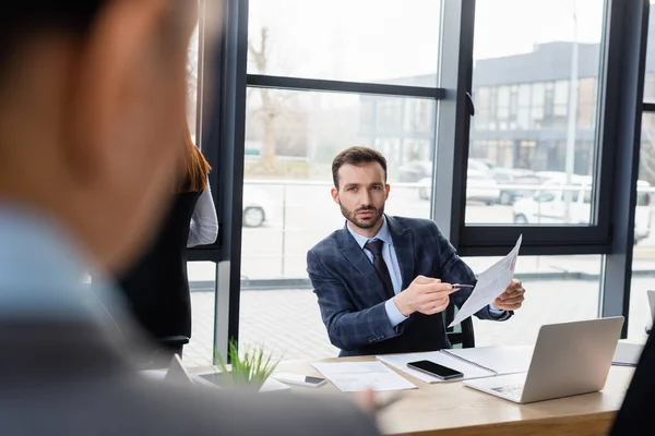 Homme d'affaires pointant vers le document près de gadgets et femme d'affaires floue — Photo de stock