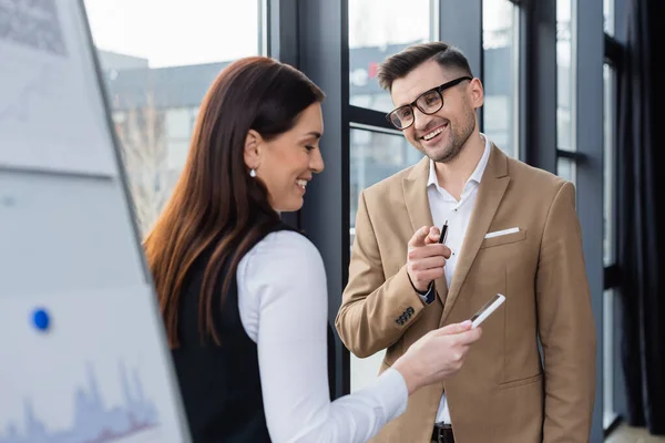 Cheerful businessman with pen pointing with finger near businesswoman using smartphone — Stock Photo