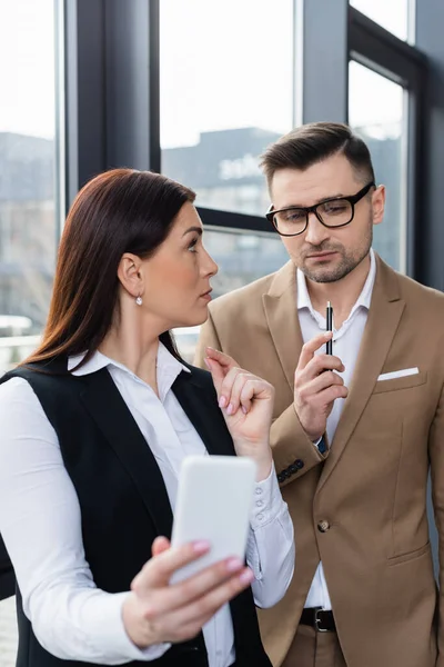 Geschäftsfrau mit verschwommenem Smartphone blickt nachdenklichen Kollegen an — Stockfoto