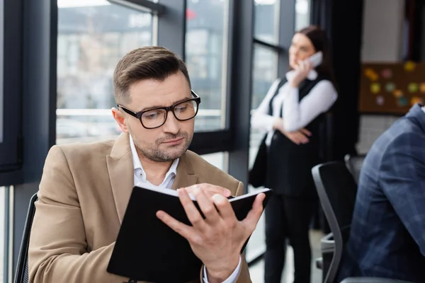 Geschäftsmann schreibt auf Notizbuch neben verschwommenen Kollegen im Büro — Stockfoto