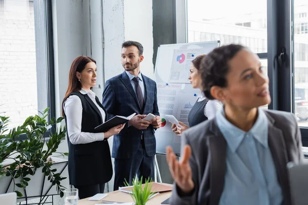 Geschäftsfrau mit Notizbuch steht neben Kollegen mit Geräten und Flipchart im Büro — Stockfoto