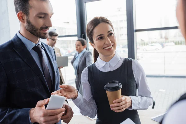 Empresária alegre com café para ir apontando para colega com smartphone — Fotografia de Stock