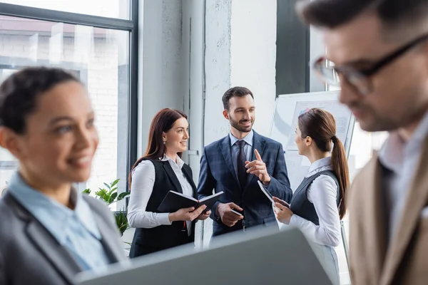 Uomo d'affari allegro che punta con il dito vicino ai colleghi con notebook e tablet digitale in ufficio — Stock Photo