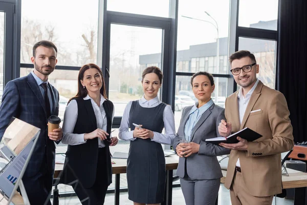 Heureux hommes d'affaires multiethniques avec café et ordinateur portable regardant la caméra dans le bureau — Photo de stock