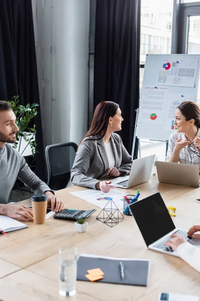 Cheerful businesswoman pointing with hand near colleagues using laptop in office — Stock Photo