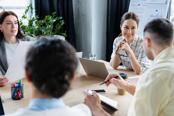 Mujeres de negocios sonrientes mirando a un colega borroso con cuaderno en la oficina — Stock Photo