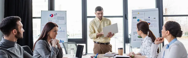 Geschäftsmann mit Laptop neben multiethnischen Kollegen und Flipcharts, Banner — Stockfoto