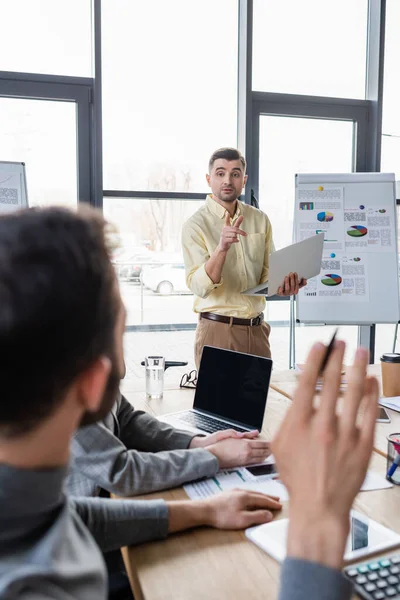 Businessman with laptop pointing at colleague on blurred foreground during meeting in office — Stock Photo