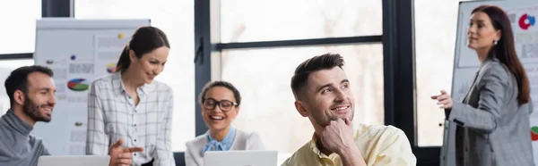 Empresario positivo mirando hacia otro lado mientras colegas multiétnicos trabajan sobre fondo borroso, pancarta — Stock Photo
