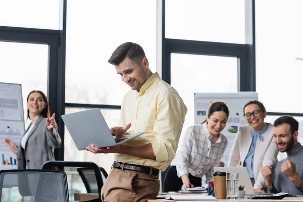 Hombre de negocios alegre usando el ordenador portátil cerca de personas de negocios multiétnicos en fondo borroso en la oficina - foto de stock