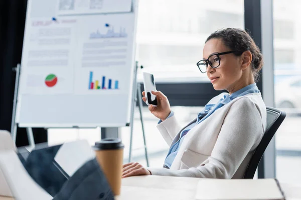 African american businesswoman with smartphone sitting near laptop and coffee to go in office — Stock Photo