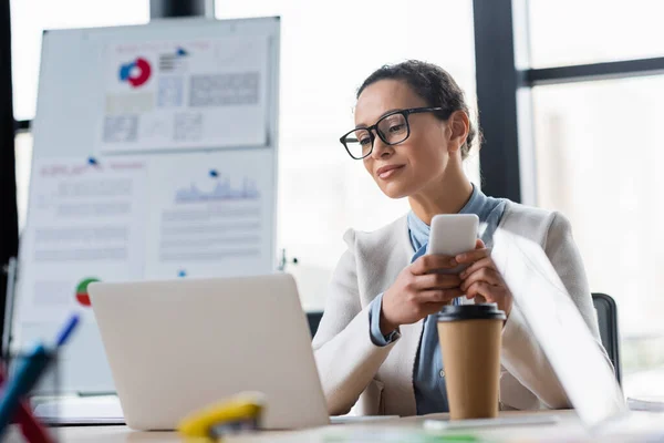 Donna d'affari afroamericana con smartphone vicino a laptop sfocato e drink da asporto in ufficio — Foto stock