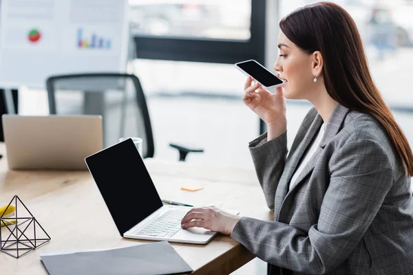 Vista lateral da mulher de negócios gravando mensagem de áudio no smartphone e usando laptop no escritório — Fotografia de Stock