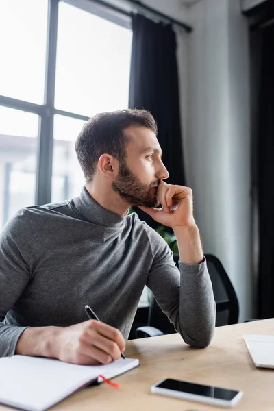 Side view of pensive manager writing on blurred notebook near smartphone — Stock Photo