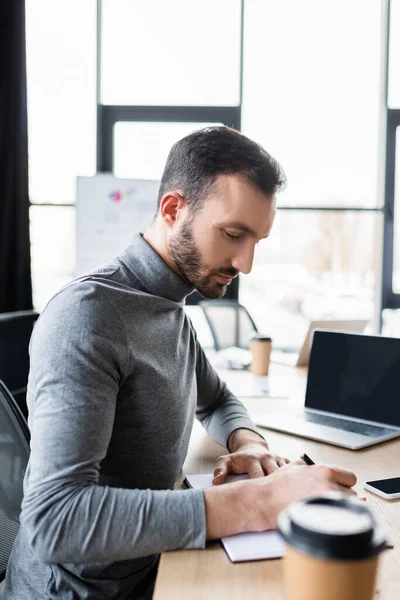 Side view of manager writing on notebook near blurred devices in office — Stock Photo