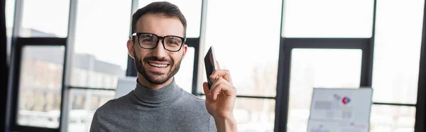 Hombre de negocios alegre con el teléfono celular mirando a la cámara en la oficina, pancarta - foto de stock