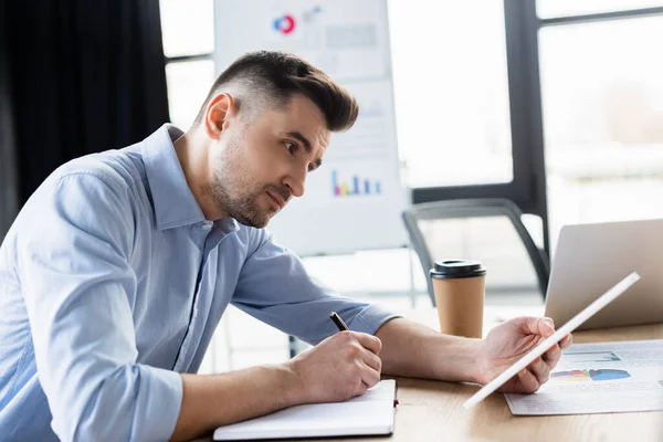 Empresario que usa tableta digital y escribe en un cuaderno cerca de una bebida para llevar - foto de stock