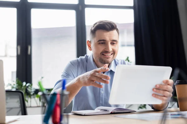 Homme d'affaires souriant utilisant une tablette numérique floue près d'un ordinateur portable et du café à emporter — Photo de stock