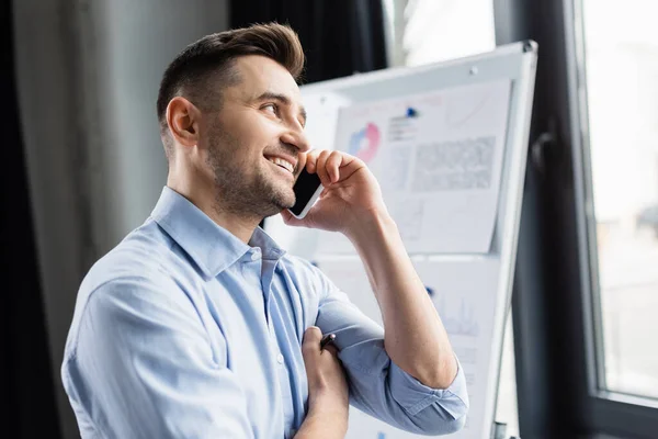 Uomo d'affari sorridente in abbigliamento formale che parla sul telefono cellulare vicino a flipchart offuscata — Foto stock